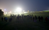 Long shot of silhouetted people walking across a field at night toward a light rig