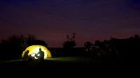 Silhouette of two campers in front of lit up tent, bikes to one side.
